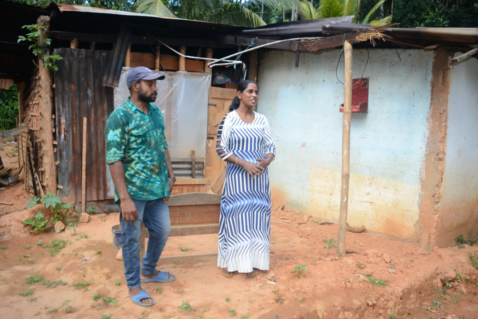 BBC/ Ranjan Arun Prasad Ravichandran, wearing a cap, green shirt, jeans and blue slip on shoes, stands next to his wife Indrani, wearing a white dress with blue lines, stand outside a building partly made from corrugated iron