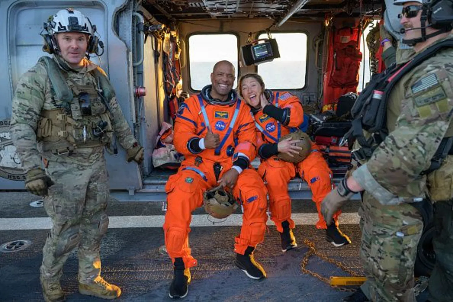 NASA The two Nasa astronauts Victor Glover and Christian Koch lean against each other in orange jumpsuits with big smiles flanked by camouflaged military personel. Victor gives a thumbs up.