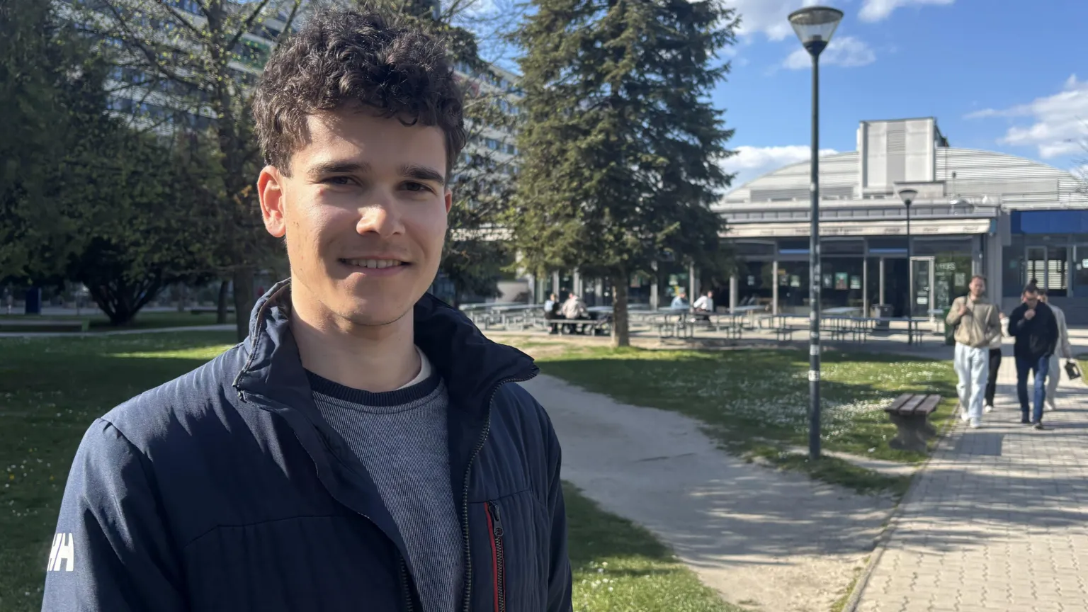 A young man with dark curly hair stands in front of a lawn and a modern building