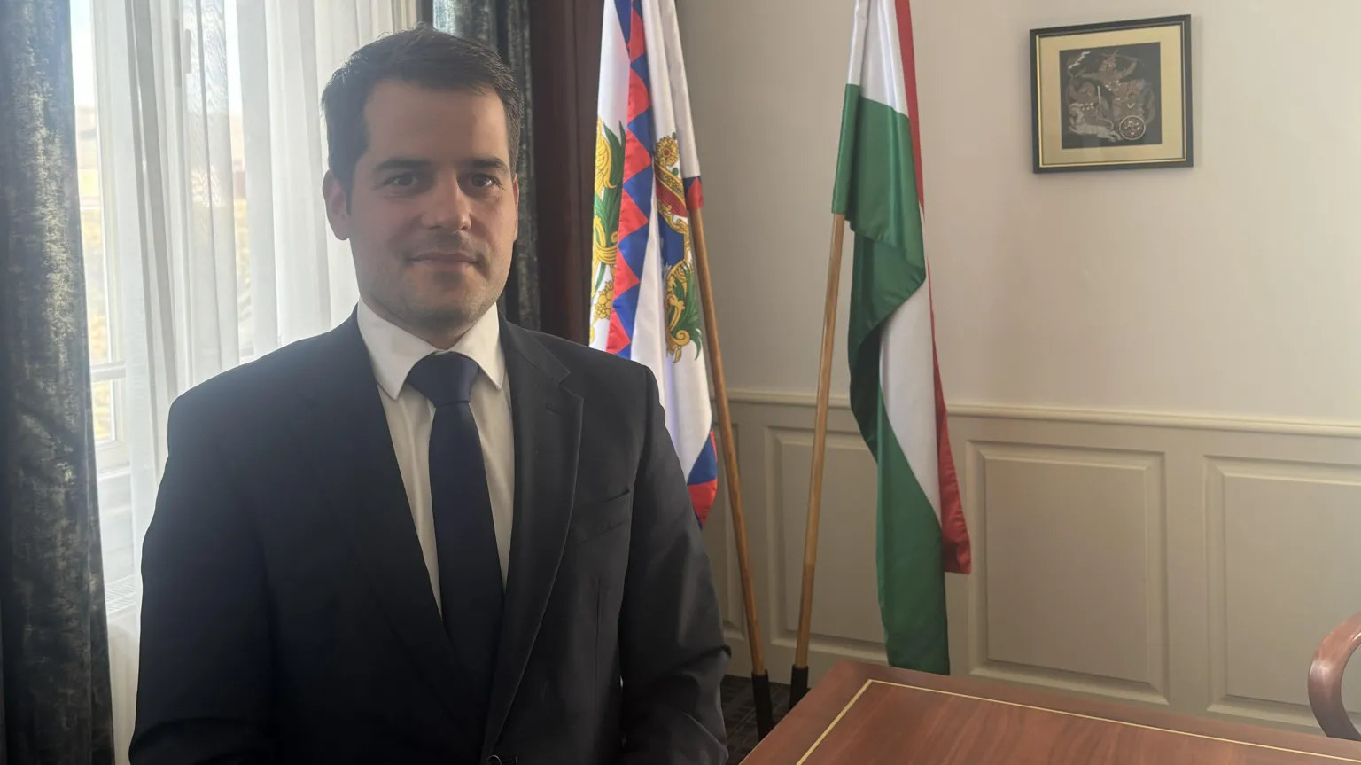 A man in a suit and tie in front of two flags