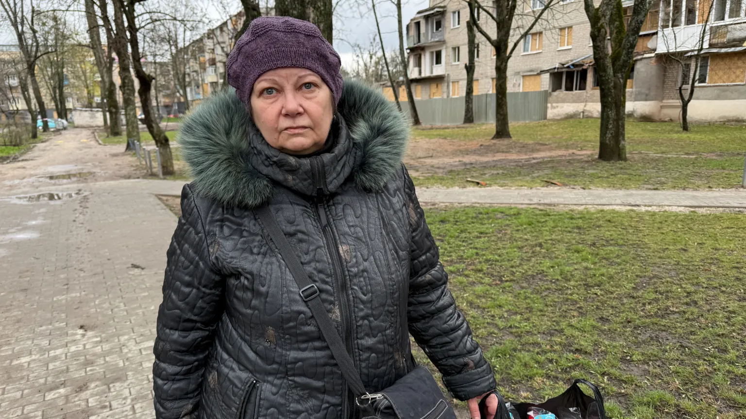 A woman in a grey coat and purple knit hat stands on the street