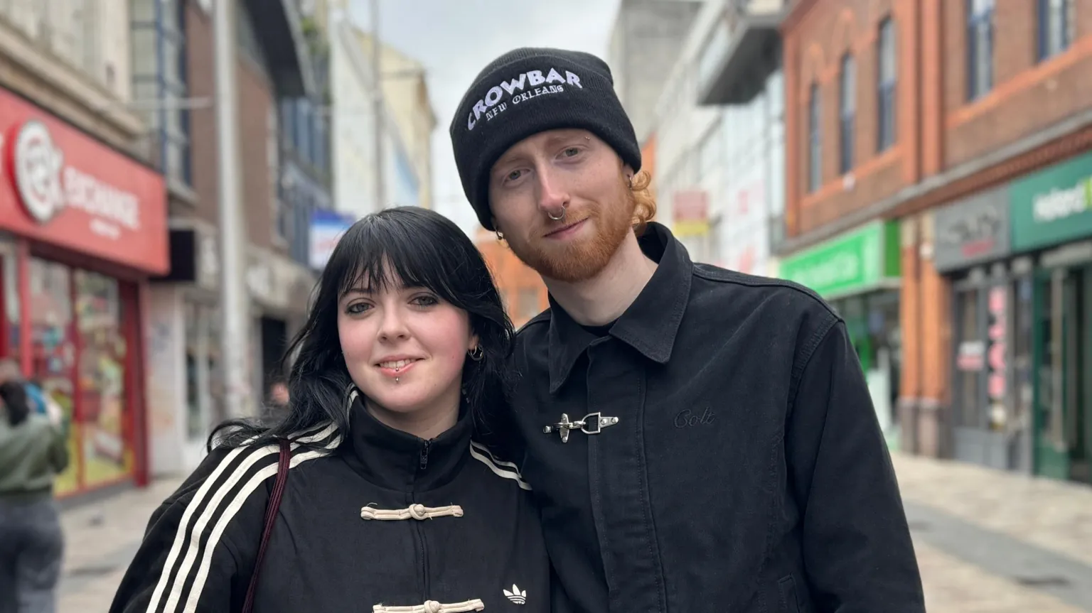 Eve and Adam standing beside each other smiling. They are both wearing black jackets. Eve has long black hair. Adam has ginger hair and is wearing a black hat.