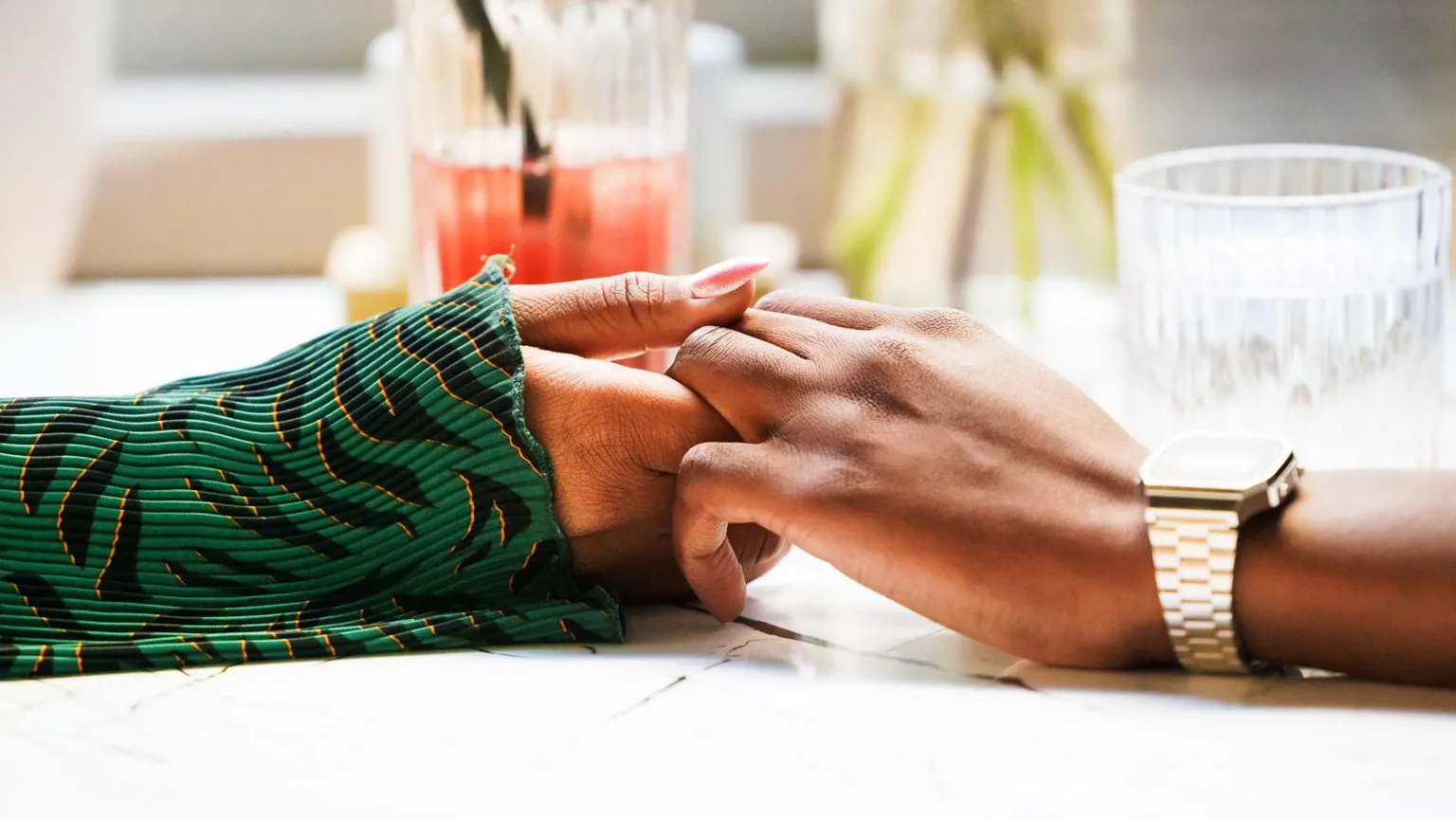 Two hands holding across a table, one has a gold watch on, the other has a green long sleeved top. There are some drinks visible in the background.
