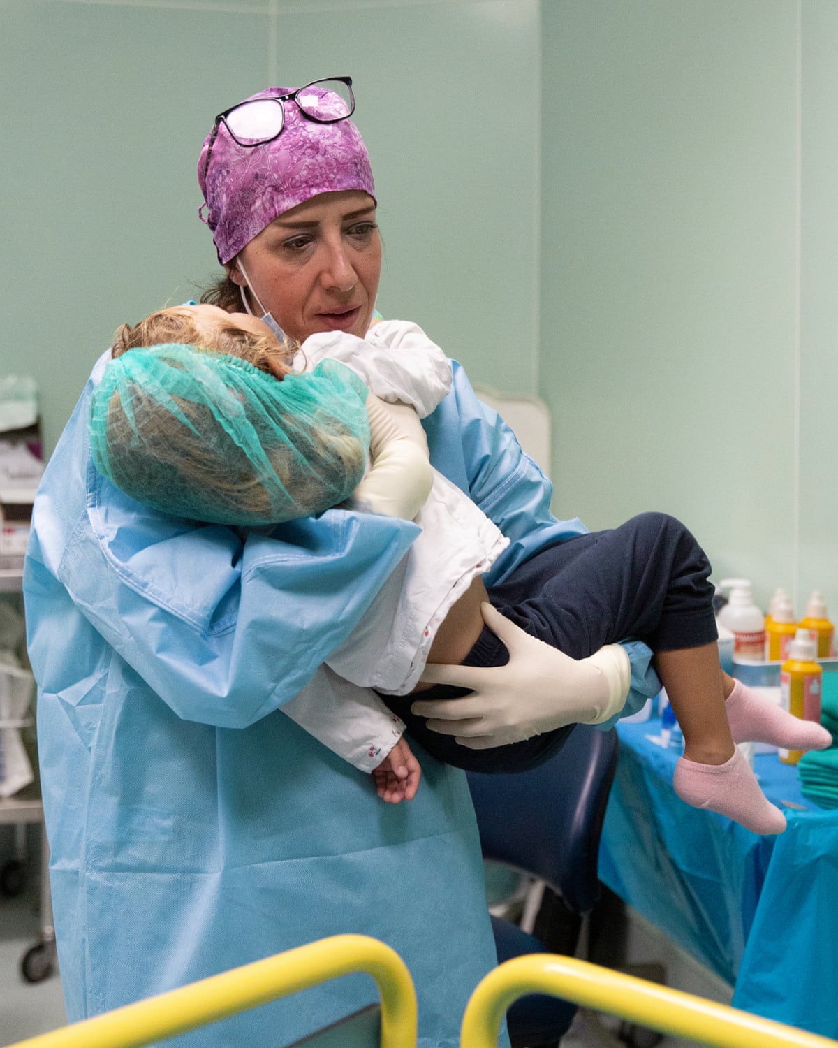 A woman in pale blue scrubs with a pink scarf over her hair holds a small girl in her arms on a hospital ward.