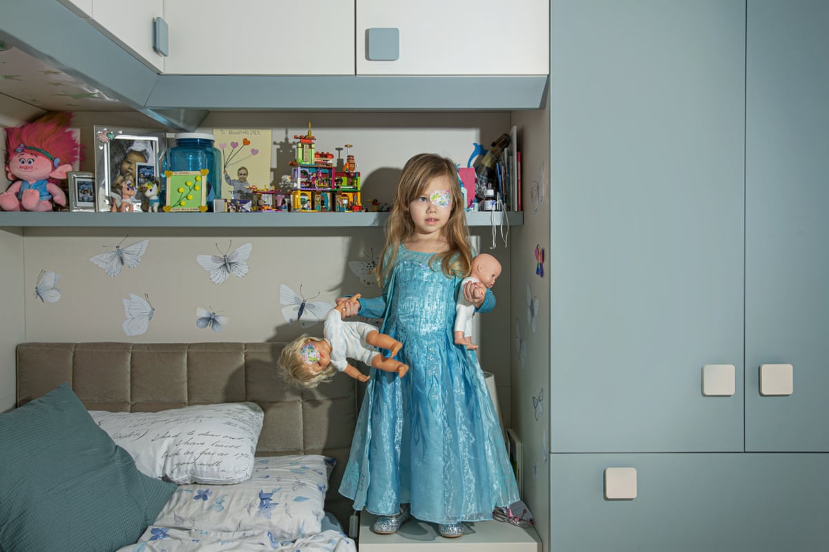 Vittoria stands on her bed in front of shelves of her toys, holding a doll in each arm. She has long blond hair and wears a shiny blue princess-style dress. She has a colourful patterned plaster over her left eye, and one of her dolls also has an eye-patch.