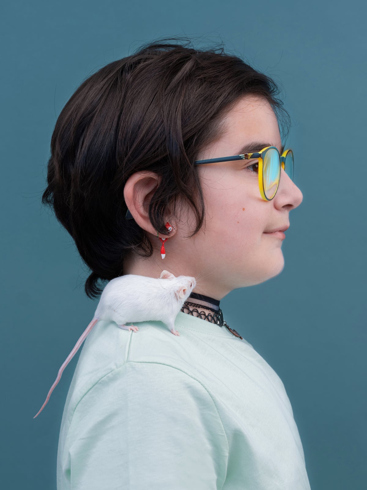 Side profile of a 10-year-old girl with ear-length dark brown hair; she wears glasses and a pale green top, and a white mouse is sitting on her shoulder.