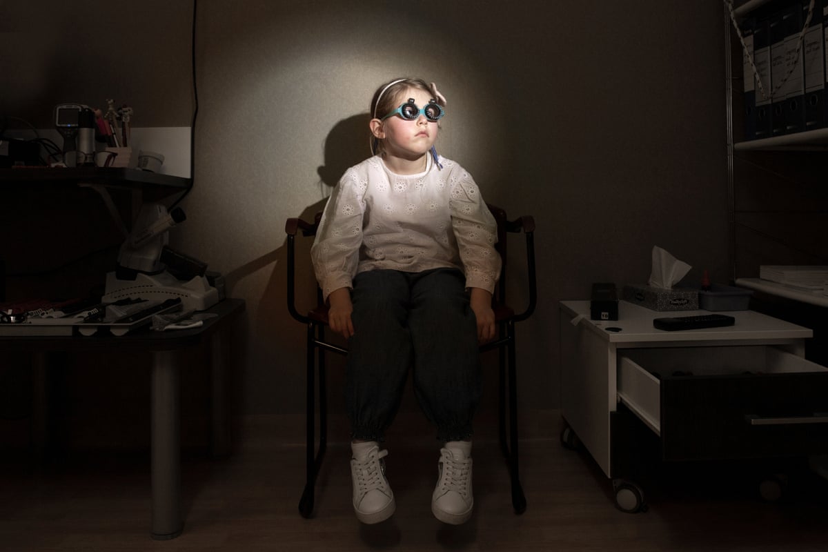 A young girl in a white top and white trainers sits in darkness at an oculist’s, with her face illuminated. She is wearing surgical glasses for tests that look like goggles.