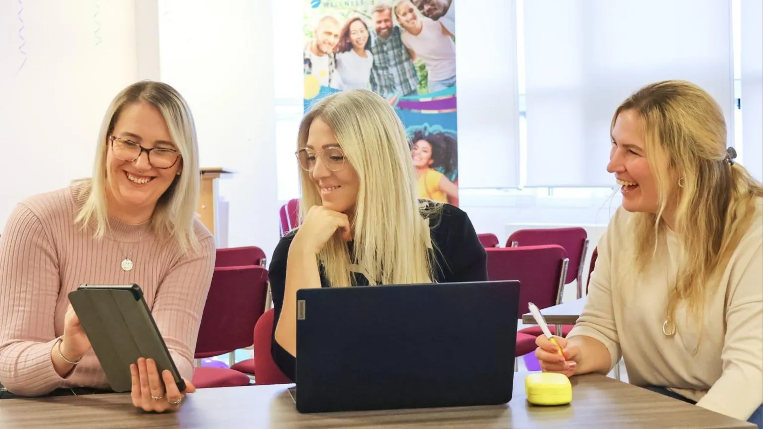 Fifty Firsts Wellness Amy has blonde hair and is wearing glasses. She is sitting behind a laptop at a table, between two women who are also blonde. They are all smiling or laughing and looking over at a tablet screen held by the woman on the left.