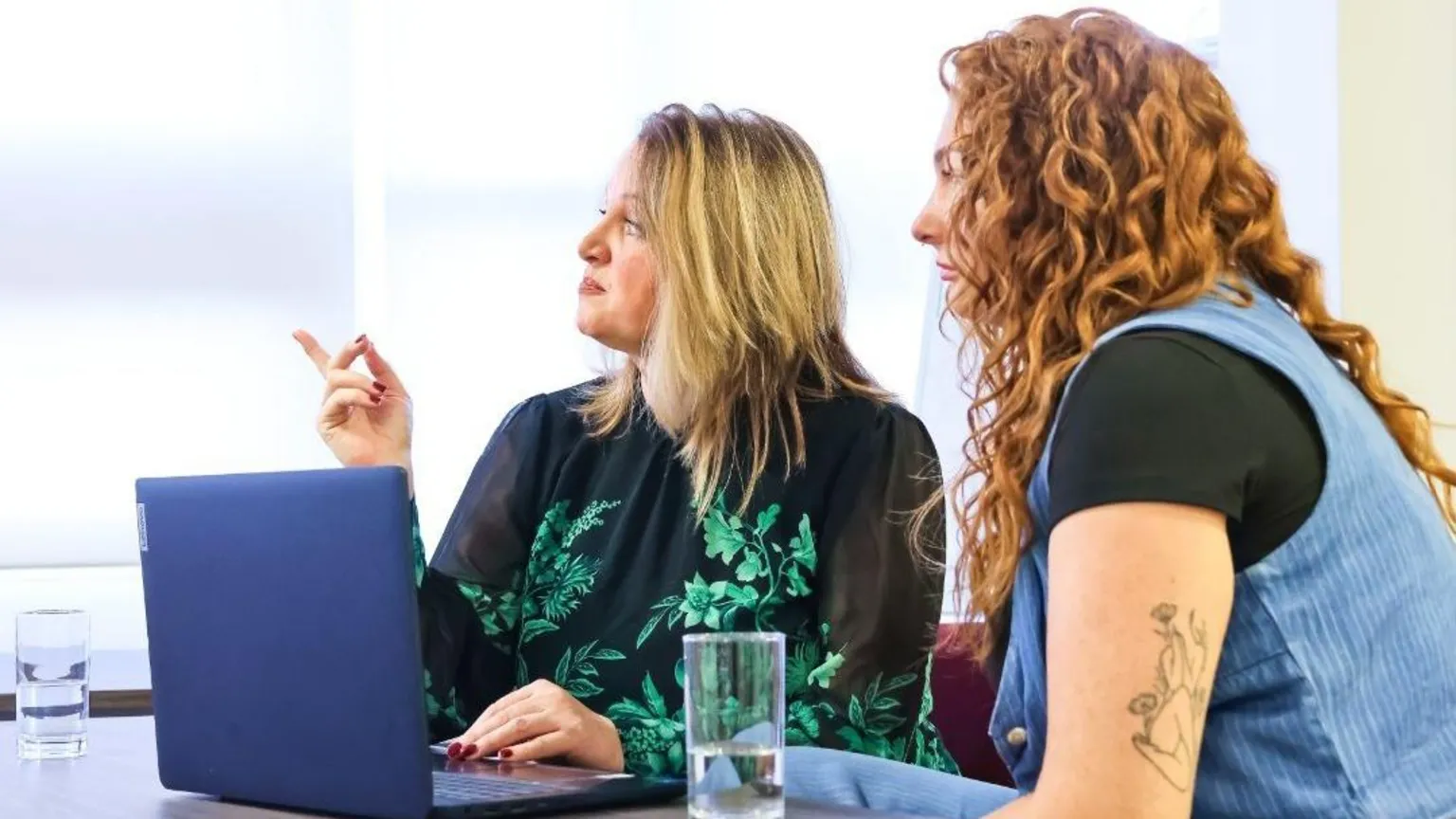 Fifty Firsts Wellbeing A blonde woman is sat at a table with a red headed woman. She has shoulder length. They have a laptop in front of them and are looking at something to the left of the camera.