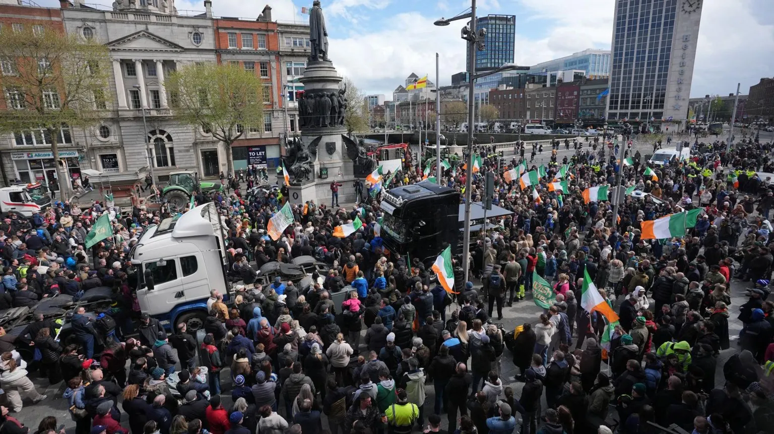 A crowd, several of them waving Irish Tricolour flags stand in O'Connell Street in Dublin. Two lorries are visible in the midst of the crowd. The statue of Daniel O'Connell can be seen in the centre of the image - bronze on top of a large stone plinth.