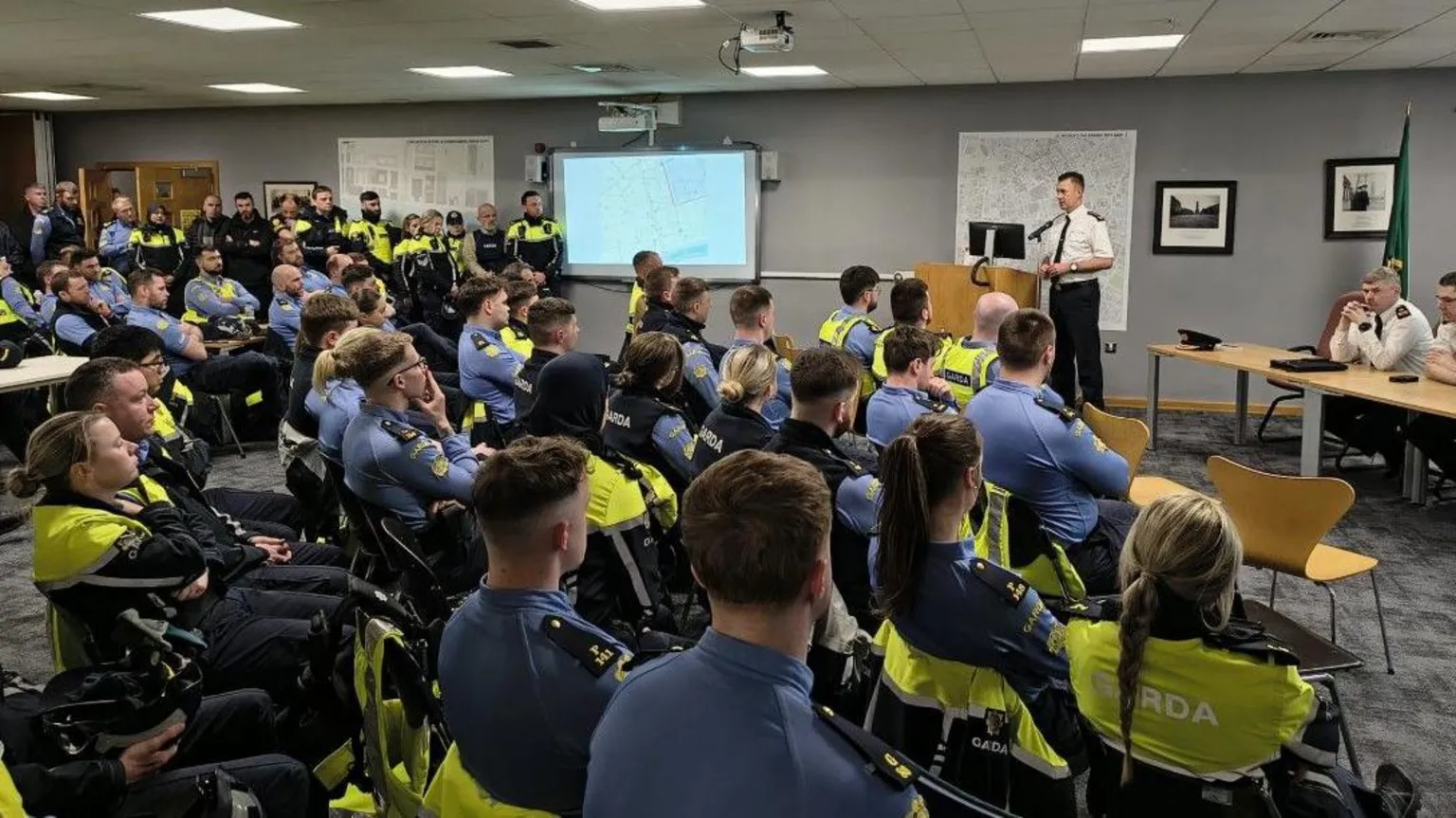 An Garda Síochána The picture shows three senior police officers in white shirts in front of 20-30 officers wearing blue shirts and high-viz jackets in a briefing room. They are listening to one of the senior officers talk, behind him is a map and there is also a map on an interactive whiteboard along the wall.
