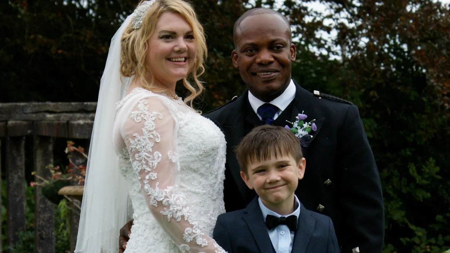Bride and groom standing outdoors with a young boy in a suit, posing for a wedding photograph.