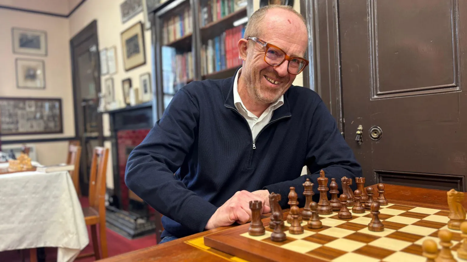 Craig Thomson sitting at a table with a chessboard in front of him. He is wearing a blue quarter zip jumper over a white shirt. He is bald and has glasses.