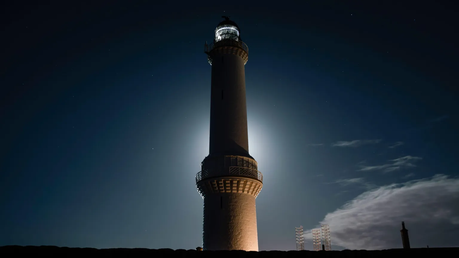 Andrew Gilchrist A tall lighthouse rises against a dark night sky, its illuminated tower glowing softly with a halo of light and faint stars overhead.