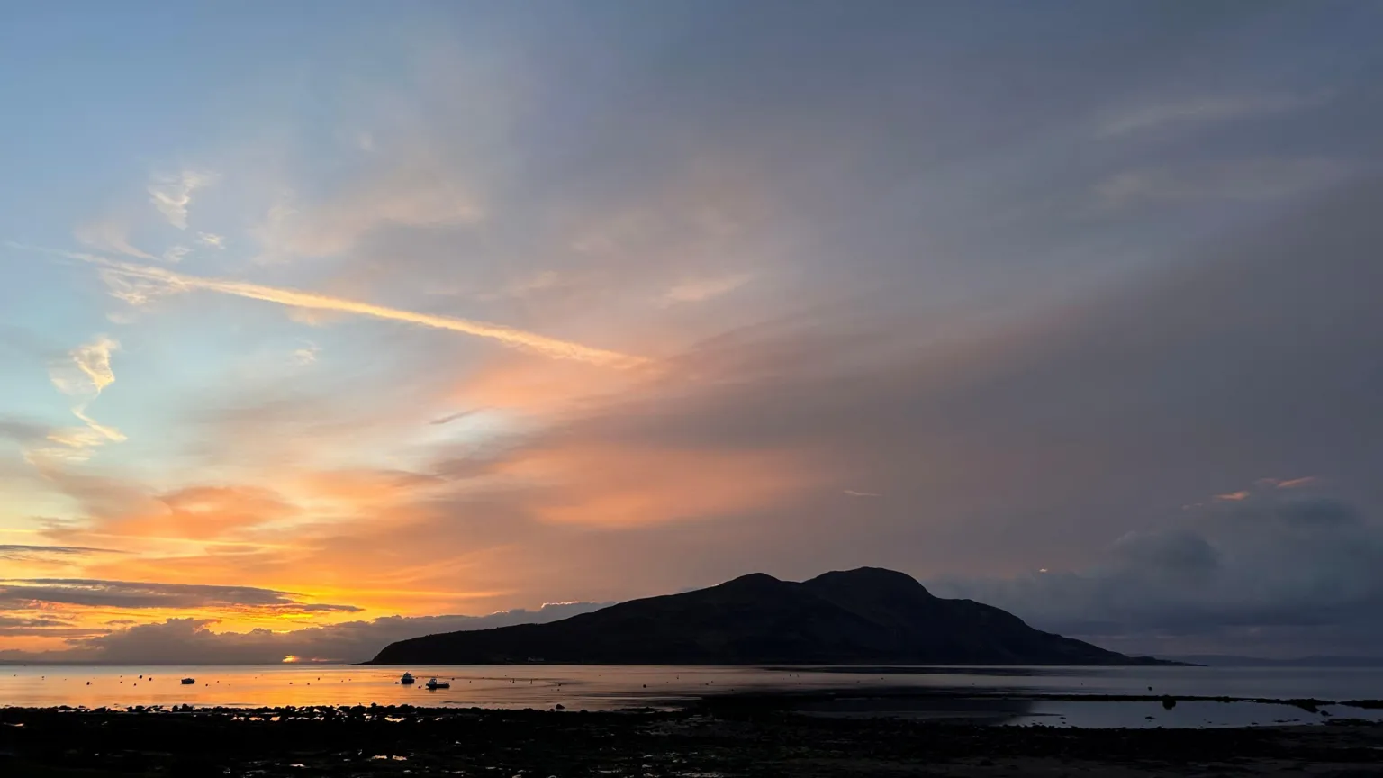 Melanie Wilde A tranquil sunset glows over a calm shoreline, with soft orange and blue clouds framing a dark, rounded island reflected in the still water.