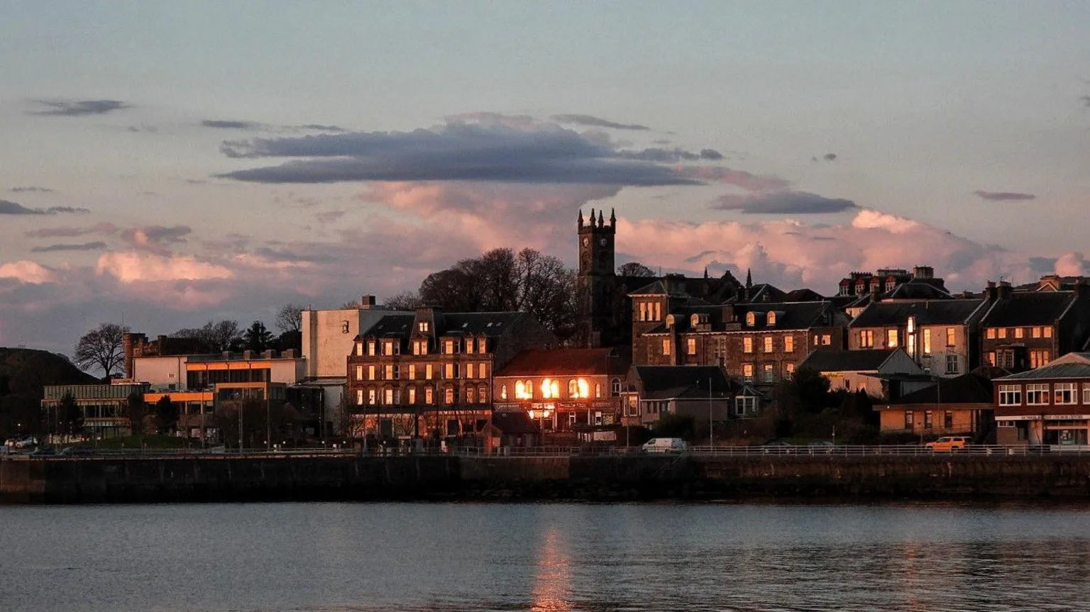 Joanna Gilpin A waterfront town at dusk, with calm water in the foreground reflecting warm lights from historic buildings along the shoreline. A church or clock tower rises above the clustered rooftops, while soft pink and grey clouds stretch across the evening sky.