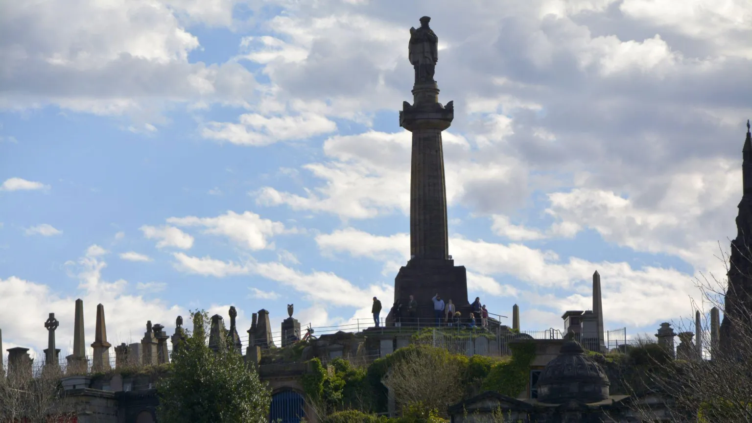 Graham Broughton A tall stone monument rises above a historic cemetery, with statues and gravestones spread across a terraced hillside. The structure stands silhouetted against a partly cloudy sky, with greenery and paths visible below.