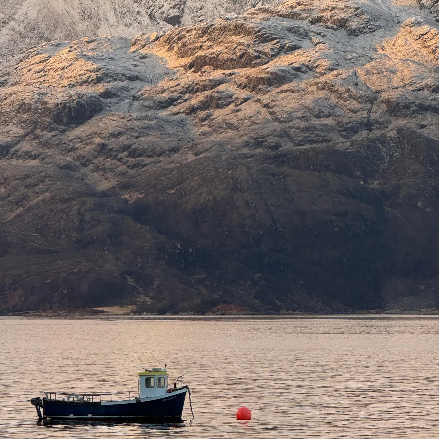 Heather McLean A small fishing boat floats on calm water in the foreground, with a red buoy nearby. Snow-dusted mountains rise steeply behind, their upper slopes catching warm light under a soft, overcast sky.