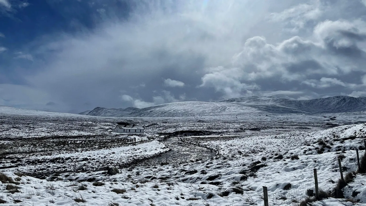 Jan Overmeer A wide, snow-covered hillside stretches into the distance beneath a dramatic, cloud-filled sky. A solitary white building sits in the middle of the landscape, with rough ground and fence posts visible in the foreground.