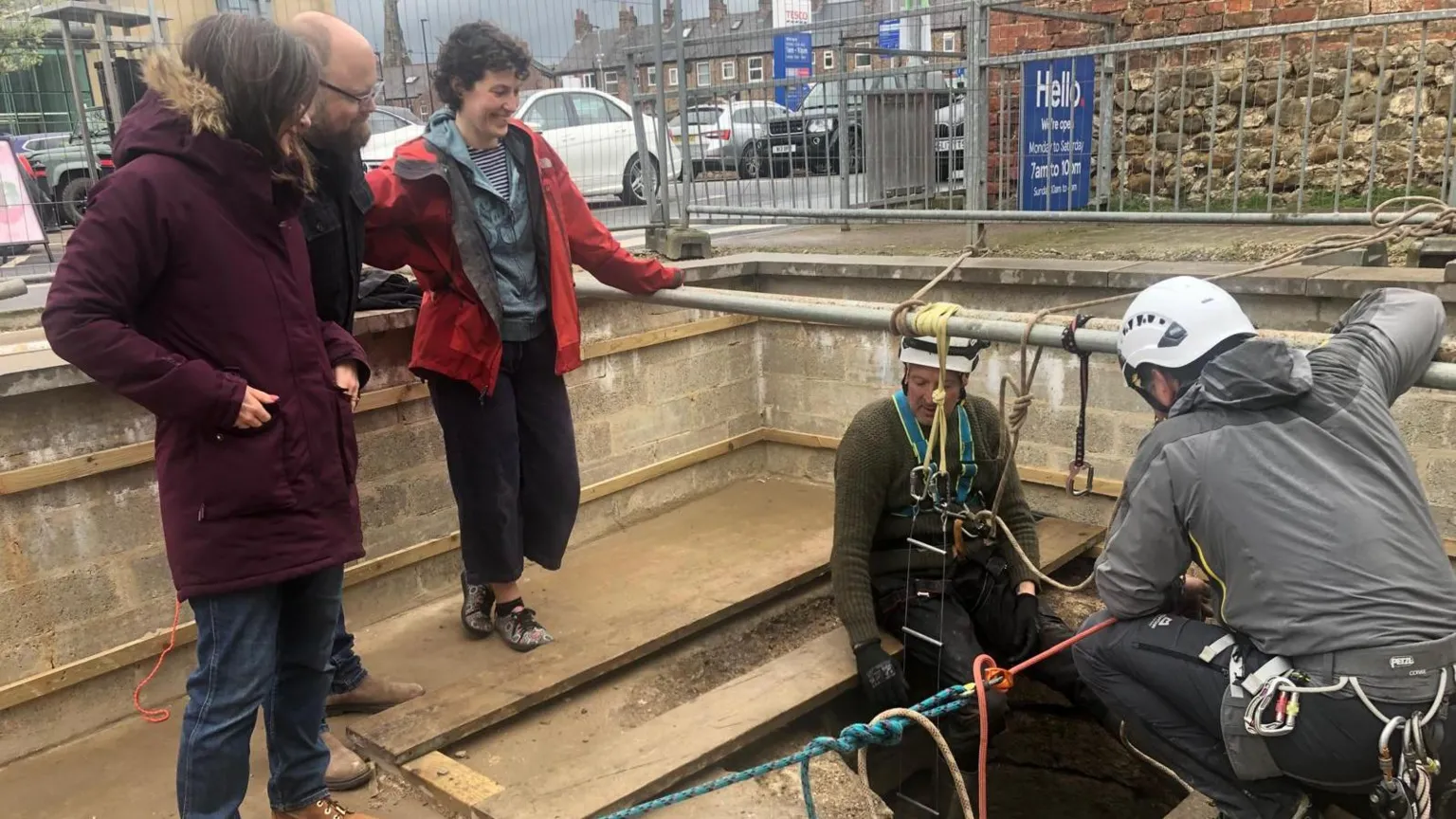 Rejoicing in Ripon Blog/Tim Flanagan A group of five people gathered around a large hole in the ground, with three standing and observing while two helmeted workers in harnesses prepare ropes and equipment for a descent.