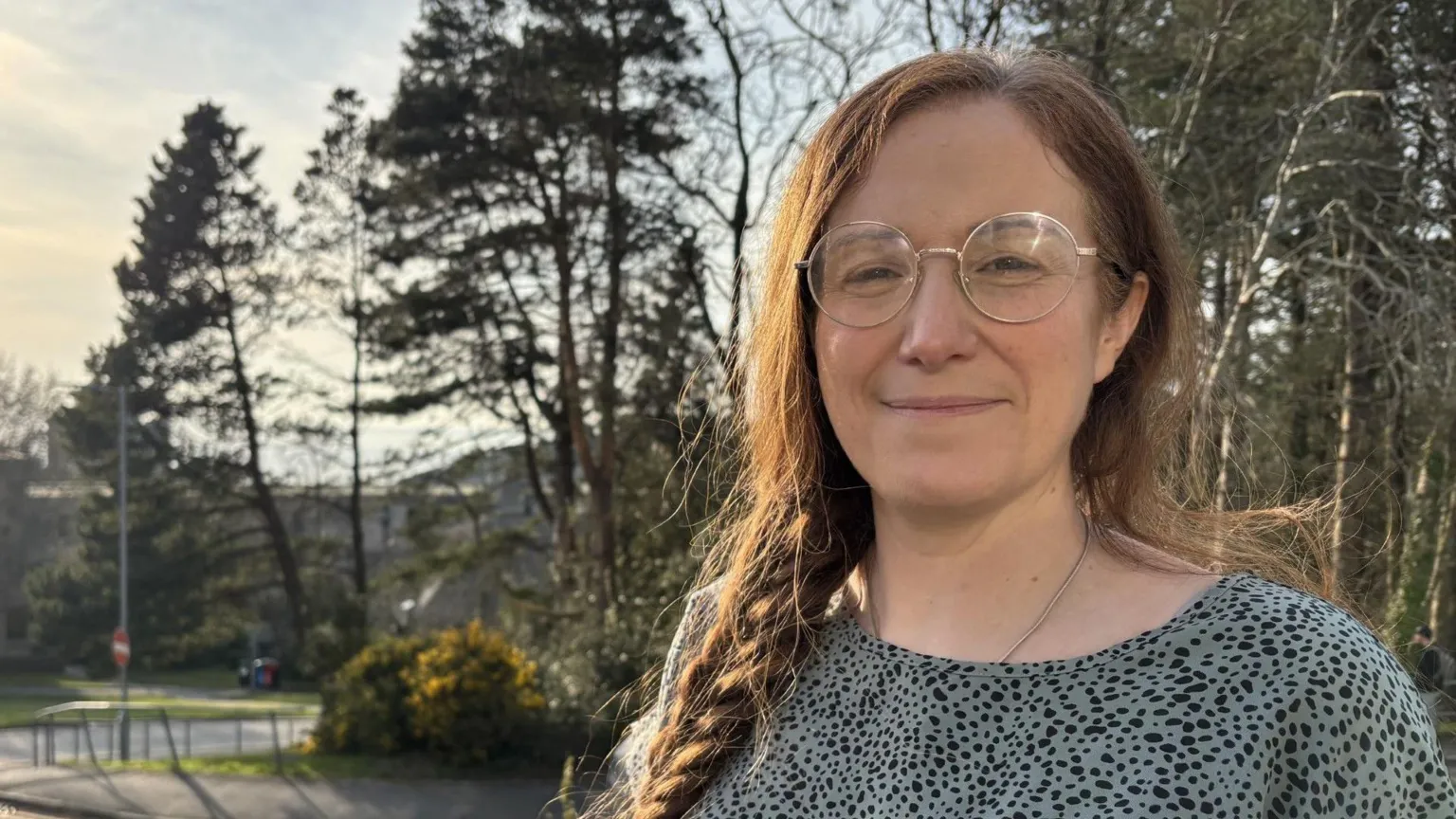 Close up of Jane Richards smiling whilst looking at the camera. She is standing in front of a road and greenery. Jane wears a blue and black spotty blouse, circular glasses and her hair is in a long side plait.