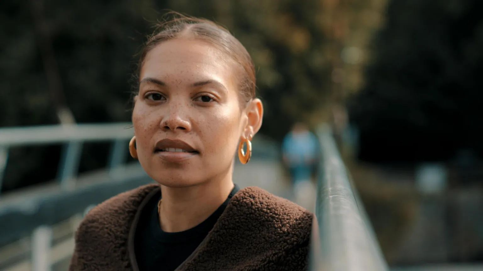 Andy Pritchard/Rondo Seren smiles at the camera. Her hair is tied back and she wears gold hoop earrings and a brown fleece top. She is standing on a bridge, but the background is blurred.