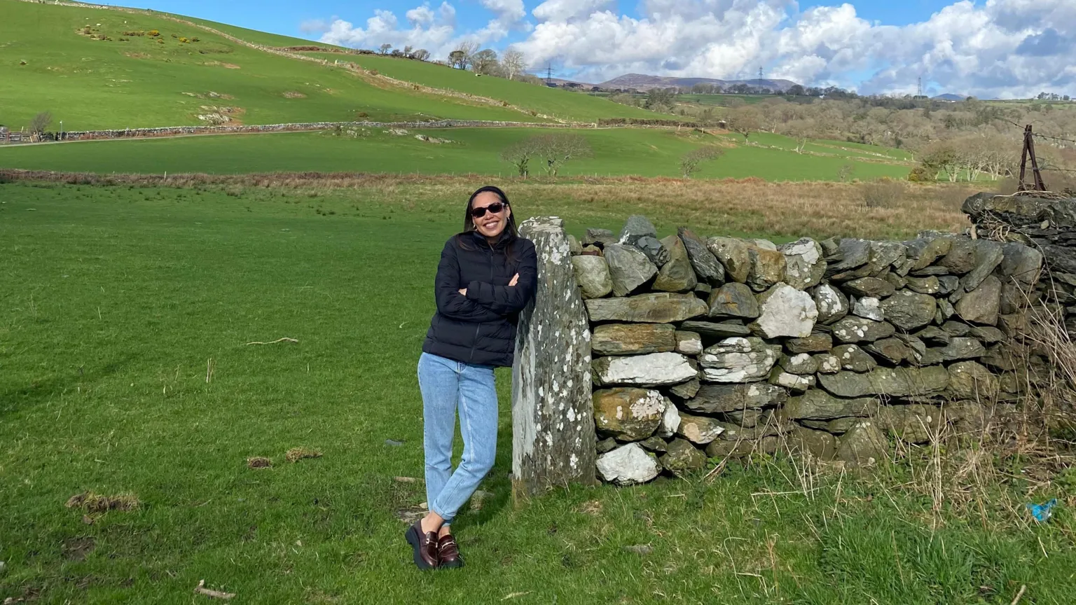 Seren Jones Seren, wearing blue jeans and a puffer jacket, leans against a stone wall in a field. She is smiling and wearing sunglasses