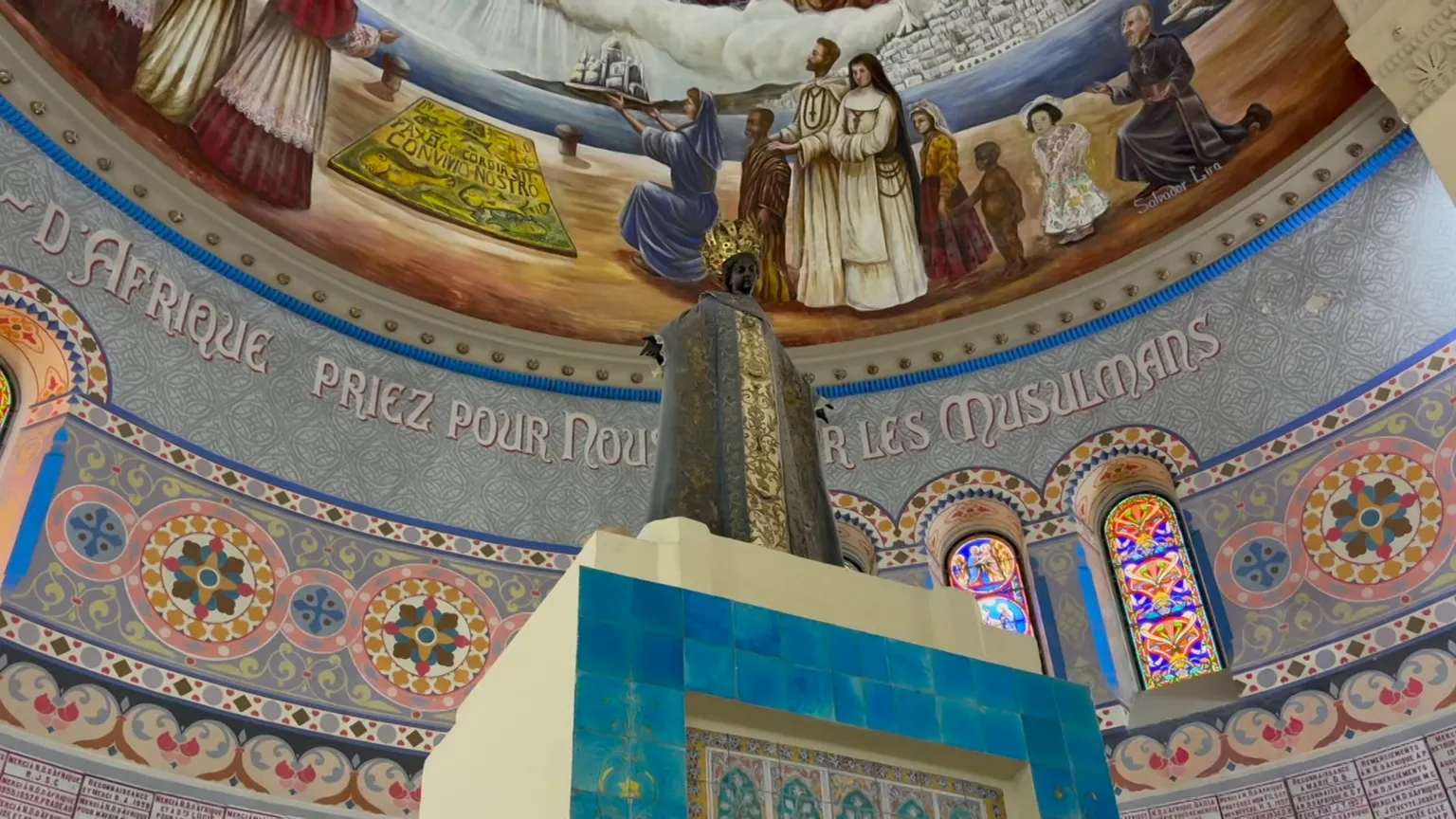 A statue of a black Madonna shot from below. Behind it is an inscription in French on a tiled wall saying, in French, 'Our Lady of Africa pray for us and the Muslims'