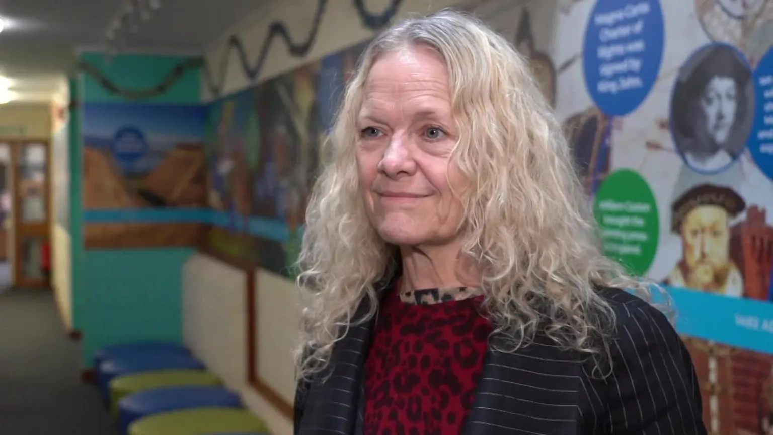 A woman with curly blonde hair standing in front of a display board in a school corridor. She is wearing a black blazer and red leopard print top.