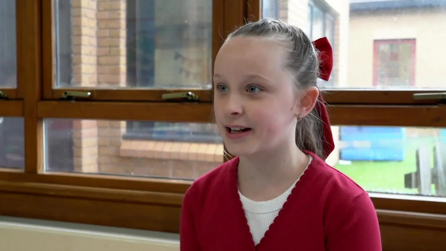 A young girl in her school uniform sitting in front of a window. She is wearing a red cardigan with a white top underneath. She is smiling and has her hair tied back with a red bow in it.