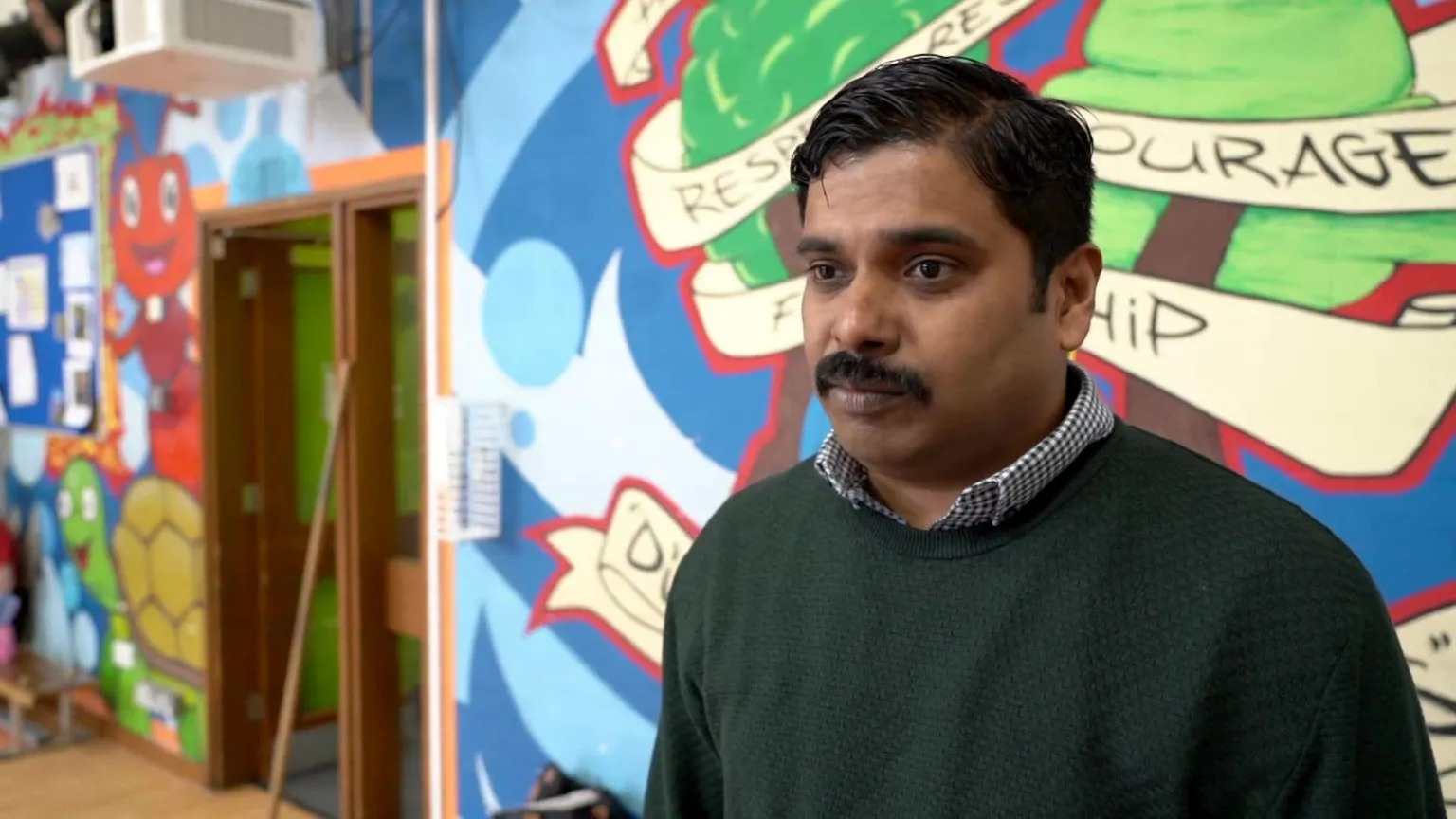 A man standing in front of a brightly painted wall in a school hall. He is wearing a green jumper with the collar of a blue check shirt underneath. He has short black hair in a side parting and a moustache.