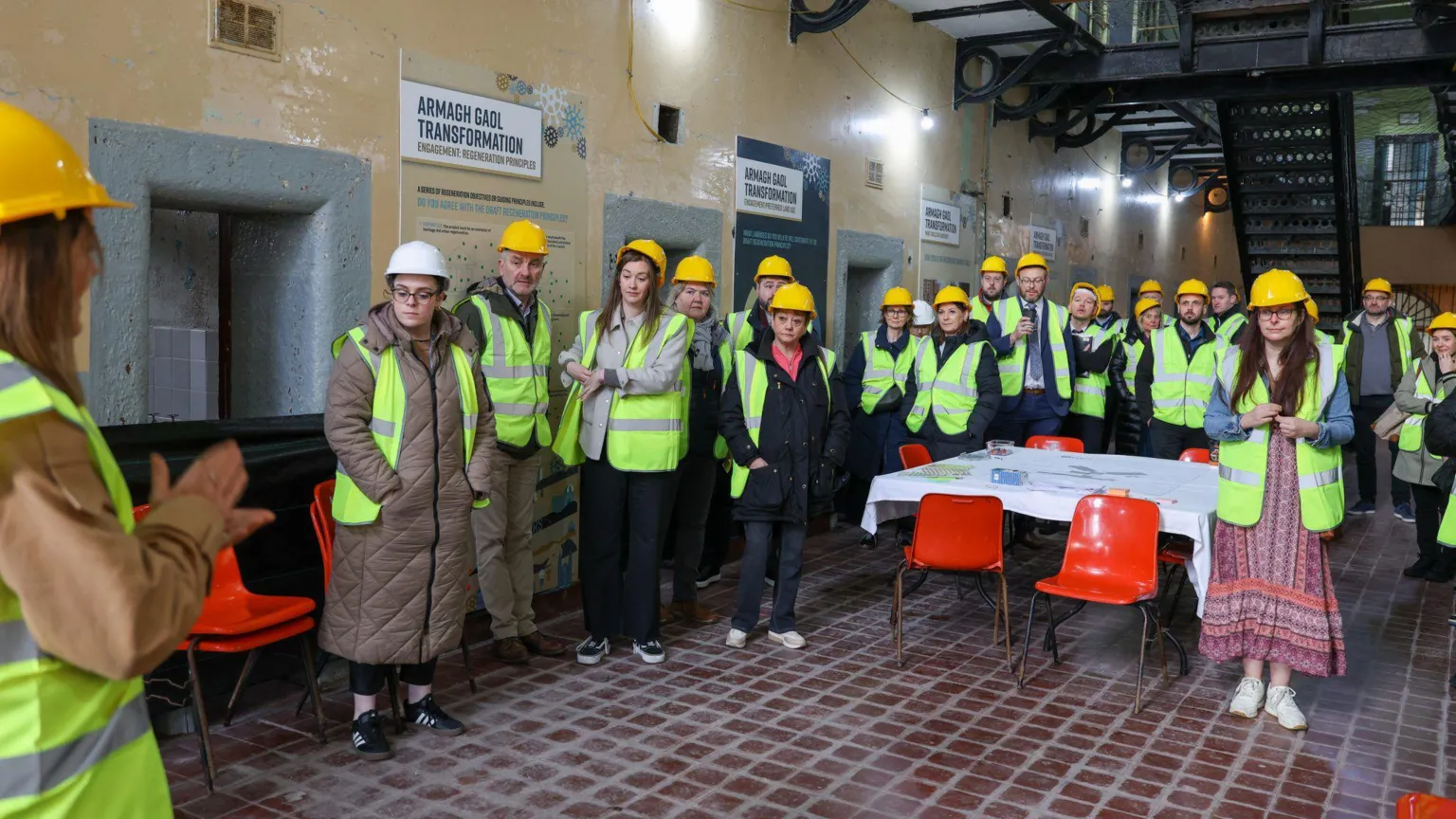Armagh, Banbridge and Craigavon Council A group of adults attend a public information session in a prison corridor inside Armagh Gaol. The visitors are all wearing yellow high-vis vests and hard hats. A woman is addressing the group, gesturing with her hands. She has long, blonde hair and is also wearing yellow protective gear. There is a black metal staircase in the background leading up to a prison landing. 