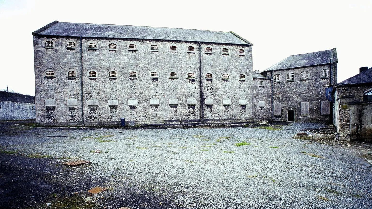 A rear view of Armagh Gaol in 2004, showing one of the two prison wings which extend diagonally into the walled prison yard. The wing is a grey, three-storey stone structure with a pitched slate roof. Each floor has 11 small arched windows. The ground floor windows are boarded up while the windows in the two upper floors are protected by prison bars. 