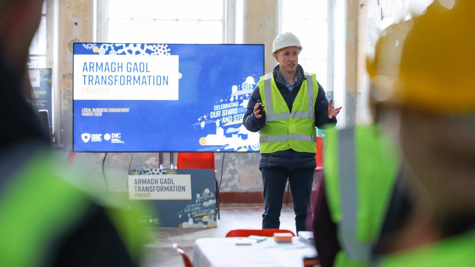 Armagh, Banbridge and Craigavon Council Kieran Carlin addressing members of the public inside Armagh Gaol during a public engagement session. He is wearing a white hard hat and a yellow high-viz vest over dark trousers and dark jacket. He is gesturing with both hands as he talks. A large TV screen behind him shows a sign which reads 