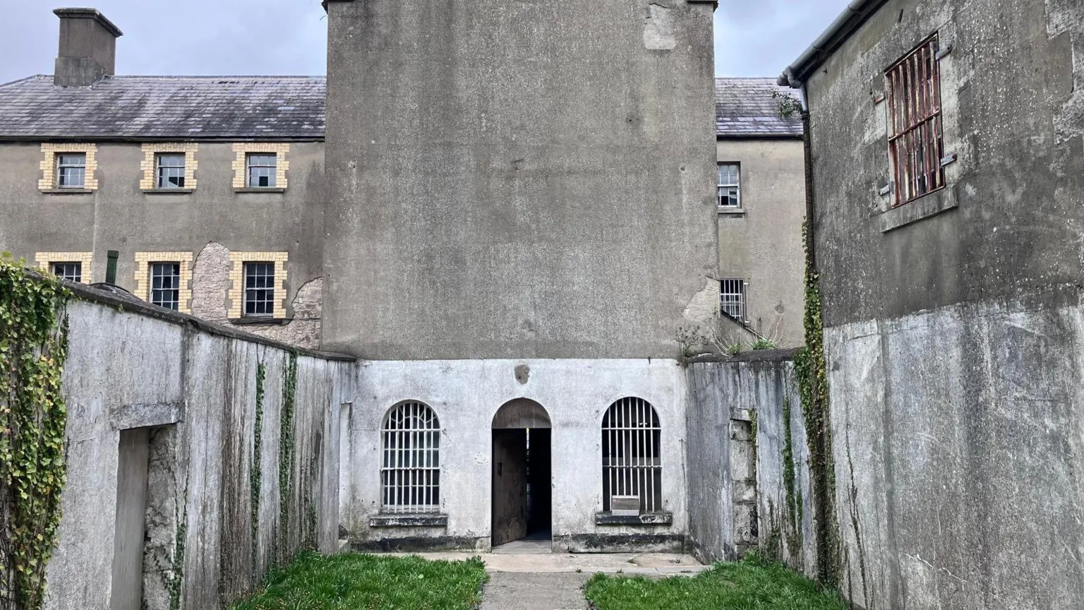 A courtyard at the rear of Armagh Gaol. A tall, plain grey building towers over a small, grassed yard, behind a larger, three-storey Georgian prison. The grey building is sealed shut apart from an arched doorway at its base, flanked by two arched windows. The windows are covered with white metal bars. Ivy is climbing up parts of the courtyard walls.