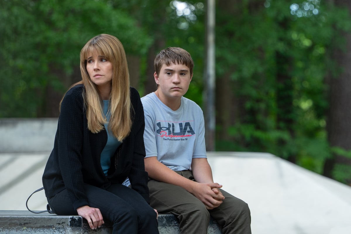 A woman with long blond hair and a fringe sits with a teenage boy both looking glum with trees in background