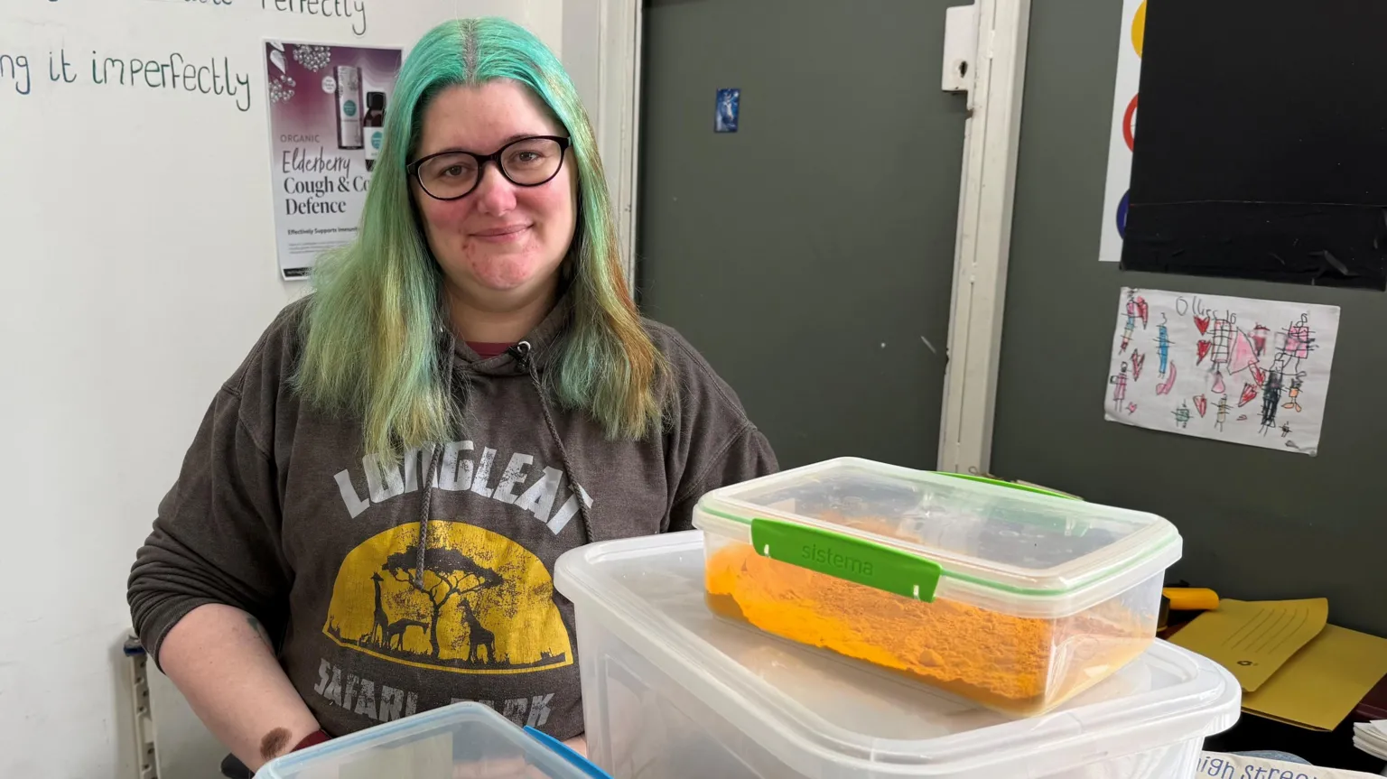 Image shows a women wearing a brown hoodie is smiling at the camera. She has blue shoulder length hair and black rimmed glasses. She is stood behind plastic containers of a yellow spice. 