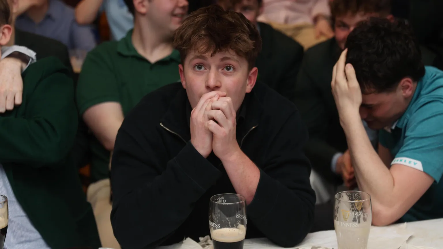 A young man wearing a black quarter-zip top is sitting with his hand clasped in front of his face watching the Masters golf intently and nervously. He is sitting at a table surrounding by other young people inside a venue. To his left is a young man with black hair with his head in his right hand.