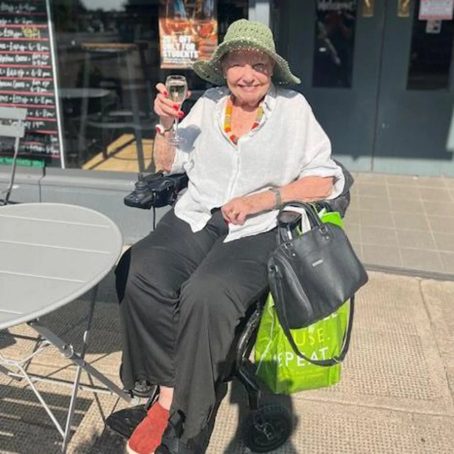 Joyce Williams A woman with a green hat holding glass of wine sits in a wheelchair at an outdoor cafe table and smiles at the camera