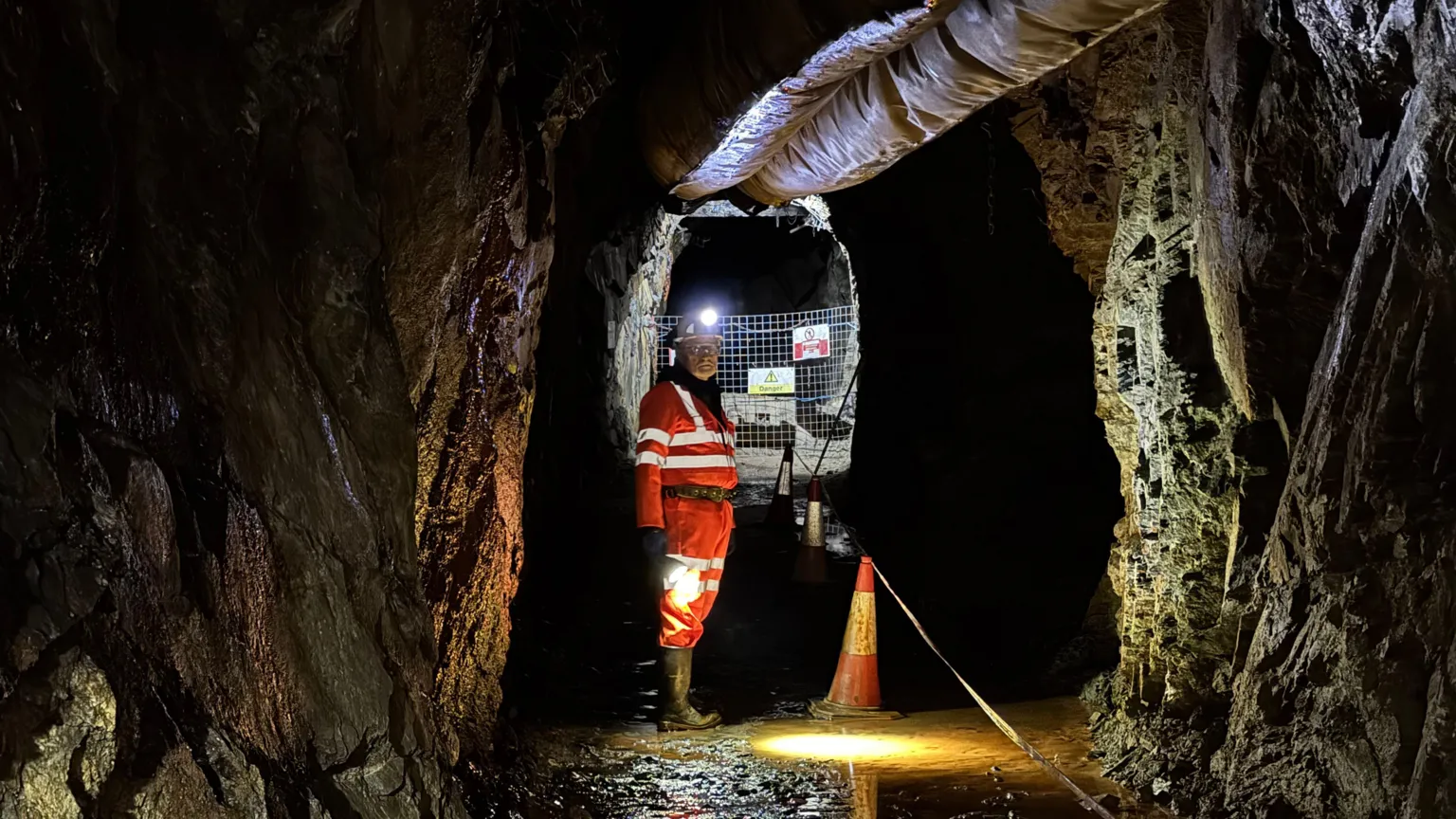 A miner dressed in orange and wearing a hard hat looks at the camera while standing in an underground mine. There is an orange and white cone beside him and his helmet lamp is reflecting off a puddle on the ground beside him.