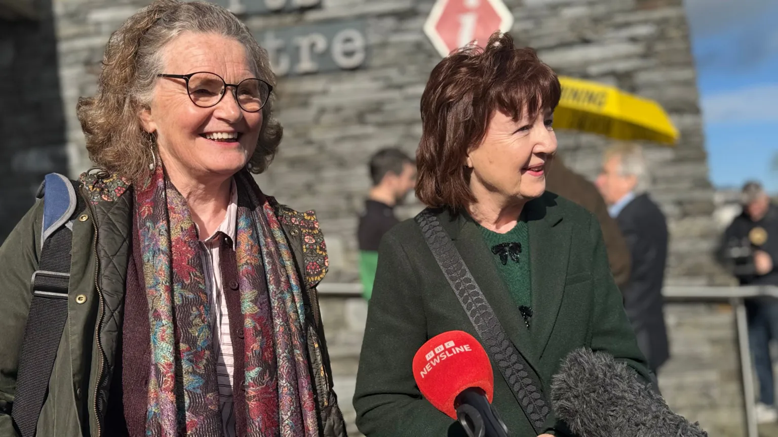 Marella Fyff has long, curly grey/brown hair and glasses while Fidema O'Kane has dark brown/red hair/ Both are standing in front of microphones