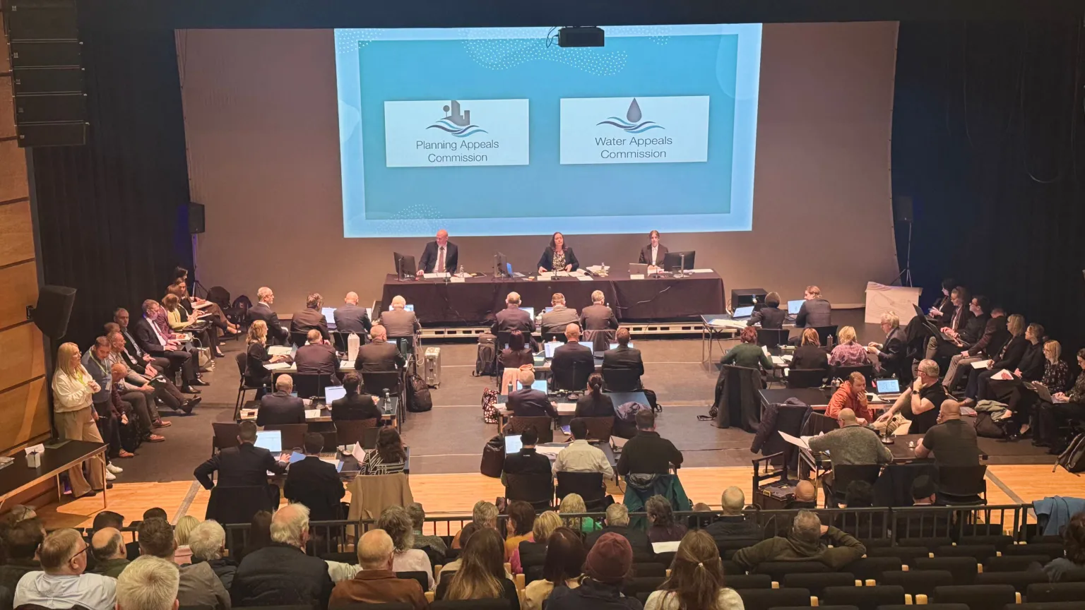 A three-person panel sits in front of a large screen in a theatre while lawyers and members of the public sit in front of them. The screen has captions on it reading