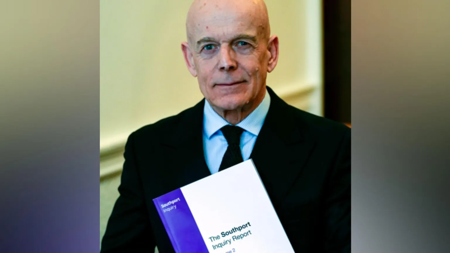  Sir Adrian Fulford, who has a bald head, is clean-shaven and wears a dark black suit, looks into the camera with a determined expression. He is holding a thick, bound copy of the Southport Inquiry Report, which has a purple and white front cover.