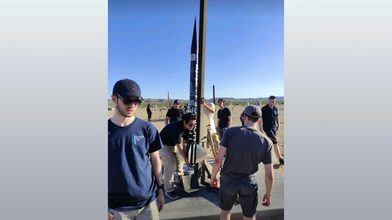 CUFSF A group of students holding a thin black rocket attached to a launch pad in a clear space on a sunny day.