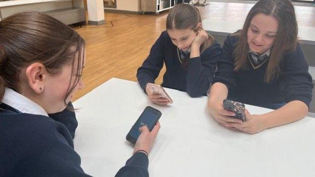 Tair disgybl yn eistedd o amgylch bwrdd mewn gwisg ysgol glas tywyll yn edrych ar eu ffonau