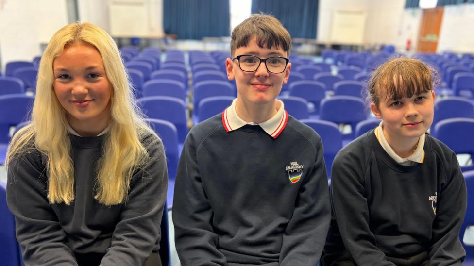 Tri disgybl - dwy ferch a bachgen yn y canol - mewn gwisg ysgol gyda siwmperi glas tywyll yn eistedd nesaf at ei gilydd mewn neuadd ysgol gyda rhesi o gadeiriau glas tu ol iddyn nhw