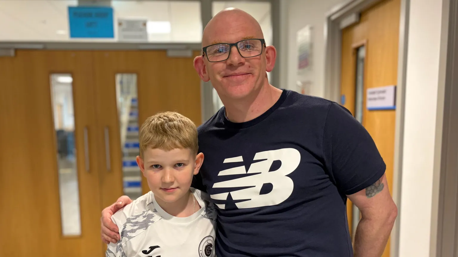 A man with a black sports t-shirt wearing glasses has his arm around a boy with blonde hair in a white football top. They are in a school corridor.