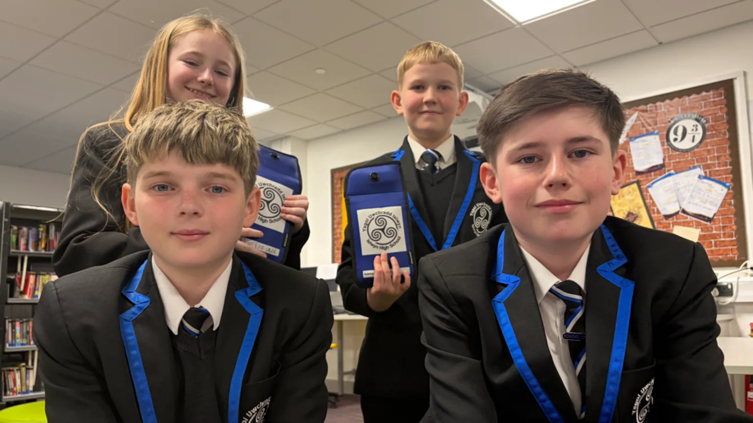 Four pupils - three boys and one girl - in school uniform wearing black blazers with blue edging. Two are sitting by a table and the two standing behind them are holding up phone pouches with the school logo on them.