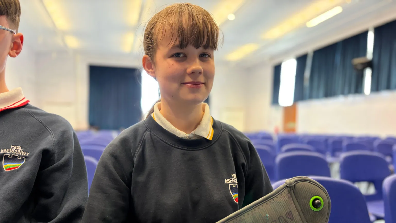 A girl in a navy school sweater with rows of blue chairs behind her, holding a pouch with the name Arwen scribbled on it