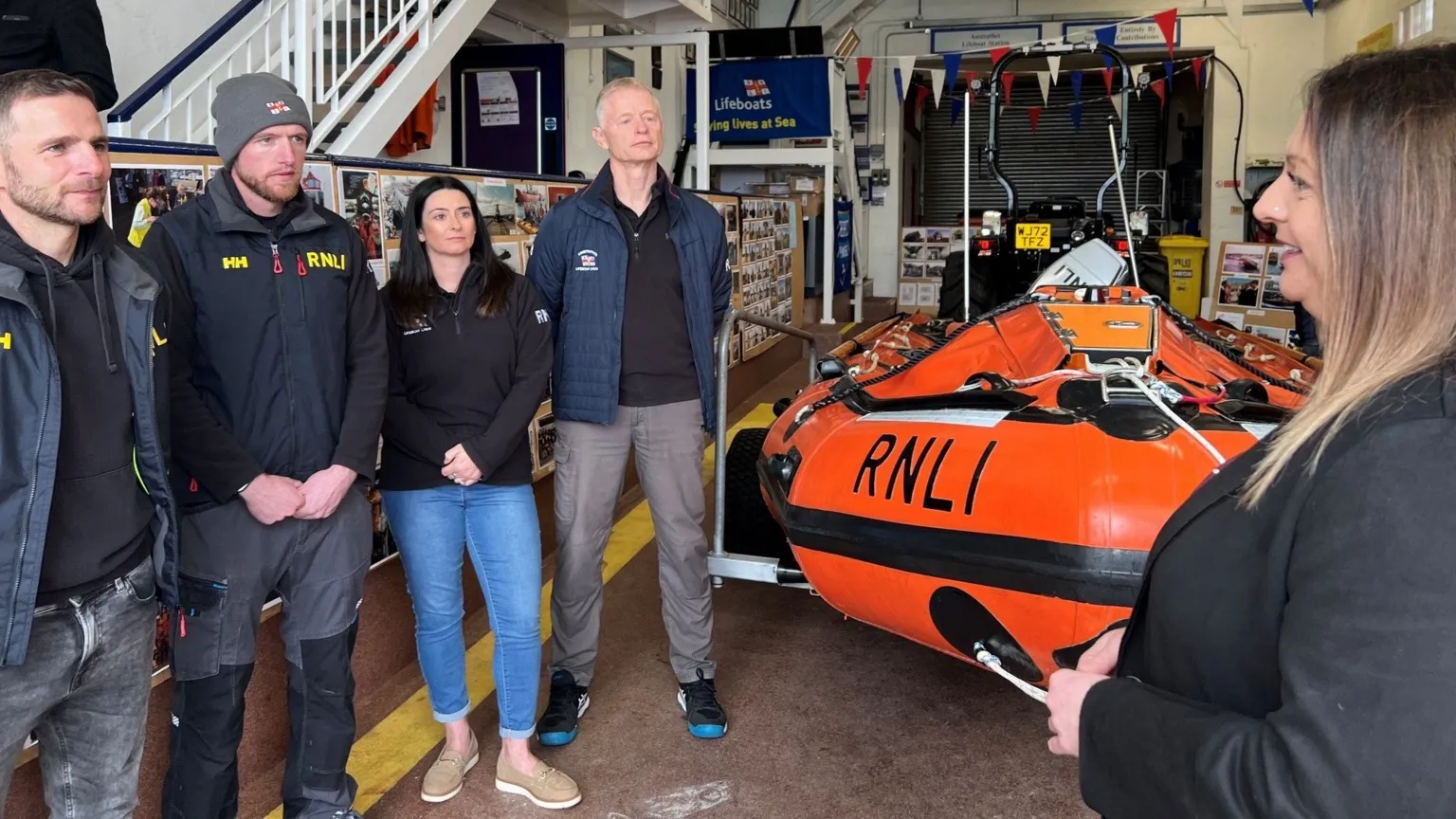 A woman speaks to three men and a woman in a lifeboat shed